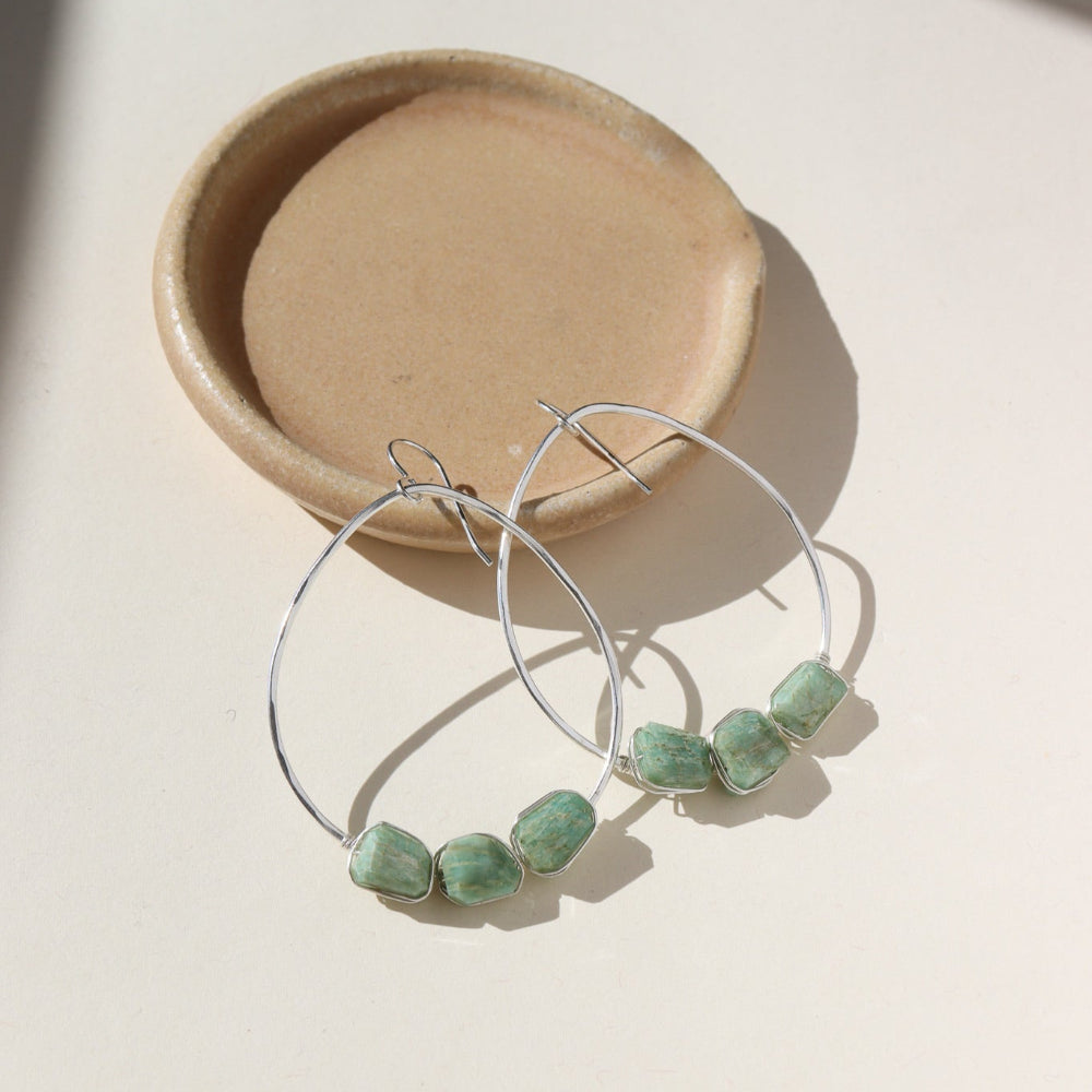 A 1.75 wide sterling silver wire hoop with three amazonite gemstones wire wrapped at the bottom of each earring. Earrings are displayed on a cream back drop. 