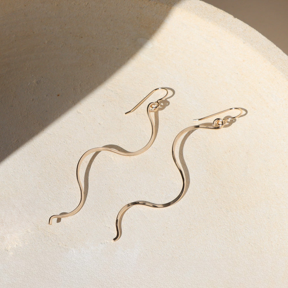 Gold snake-shaped earrings on a textured beige surface