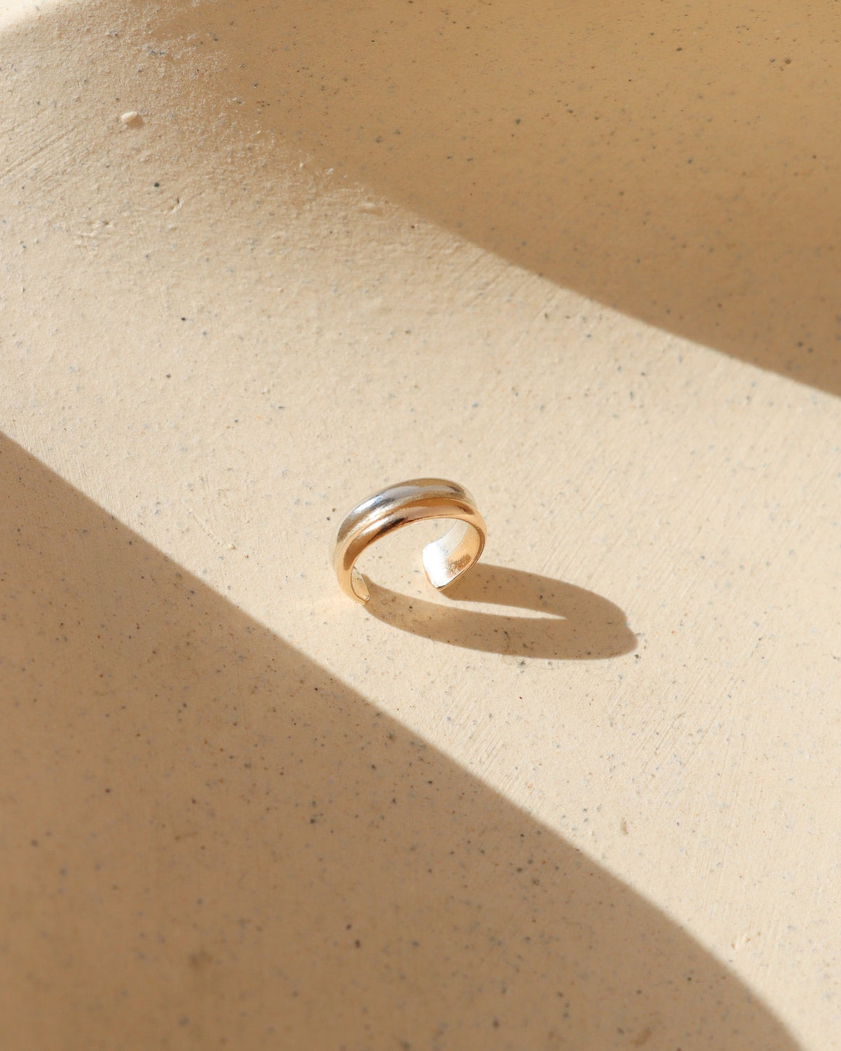 Close-up of a Token Jewelry mixed metal ear cuff featuring smooth, overlapping gold and silver bands with an open cuff design, displayed on a textured cream surface in natural sunlight.