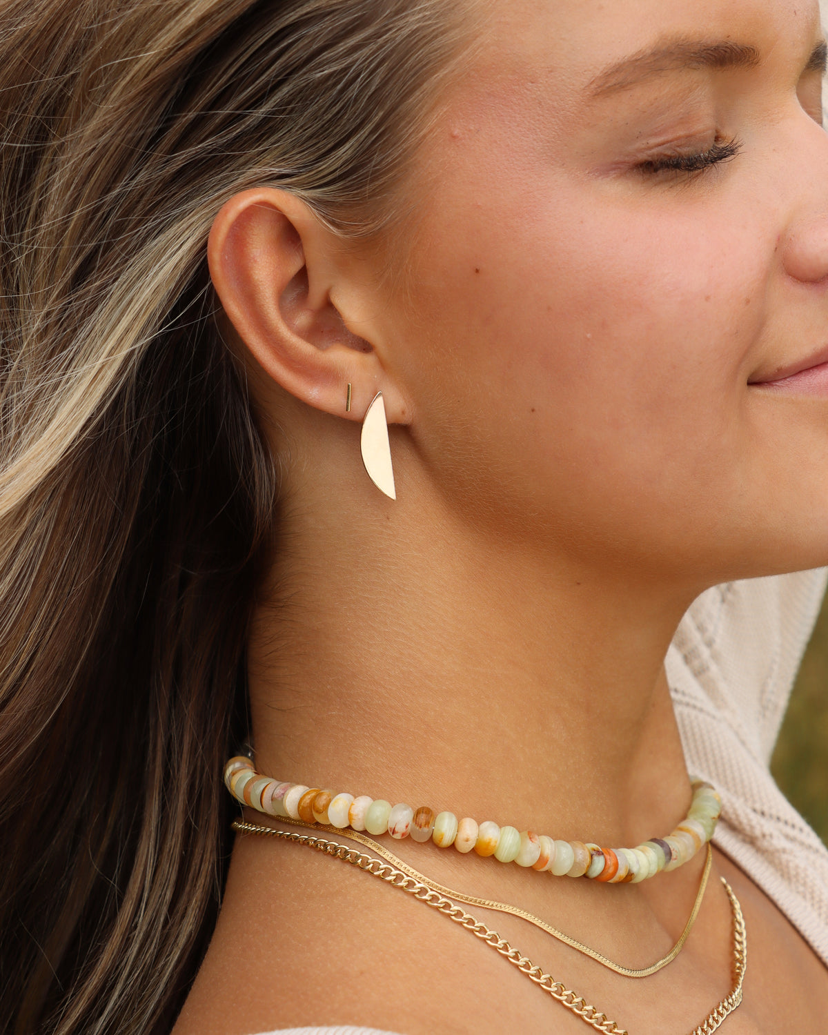 Model wearing a gold leaf-shaped statement earring with a small gold bar stud, styled with a pastel gemstone beaded necklace and layered gold chains.