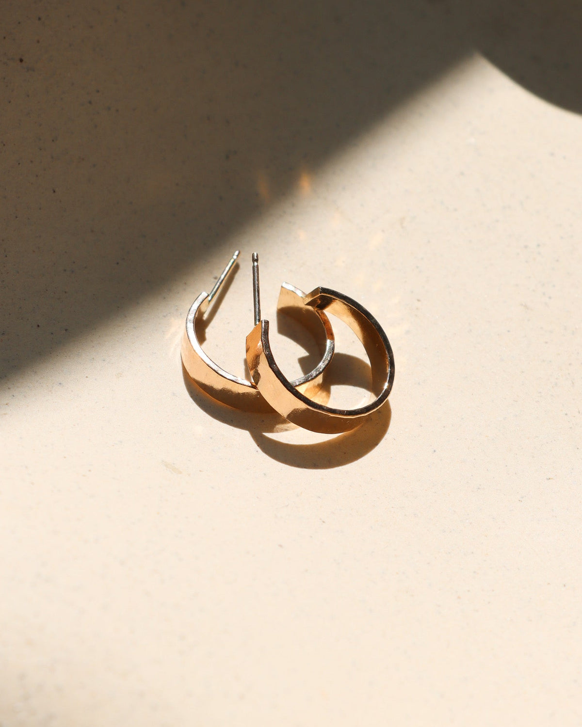 Pair of gold hoop earrings with a wide, polished design, displayed in warm sunlight on a neutral surface.