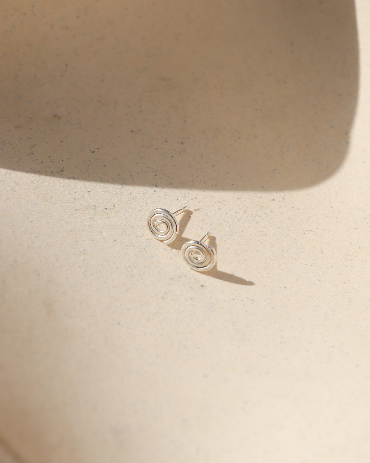Sterling silver spiral stud earrings with a delicate coiled design displayed on a textured cream surface in natural sunlight.