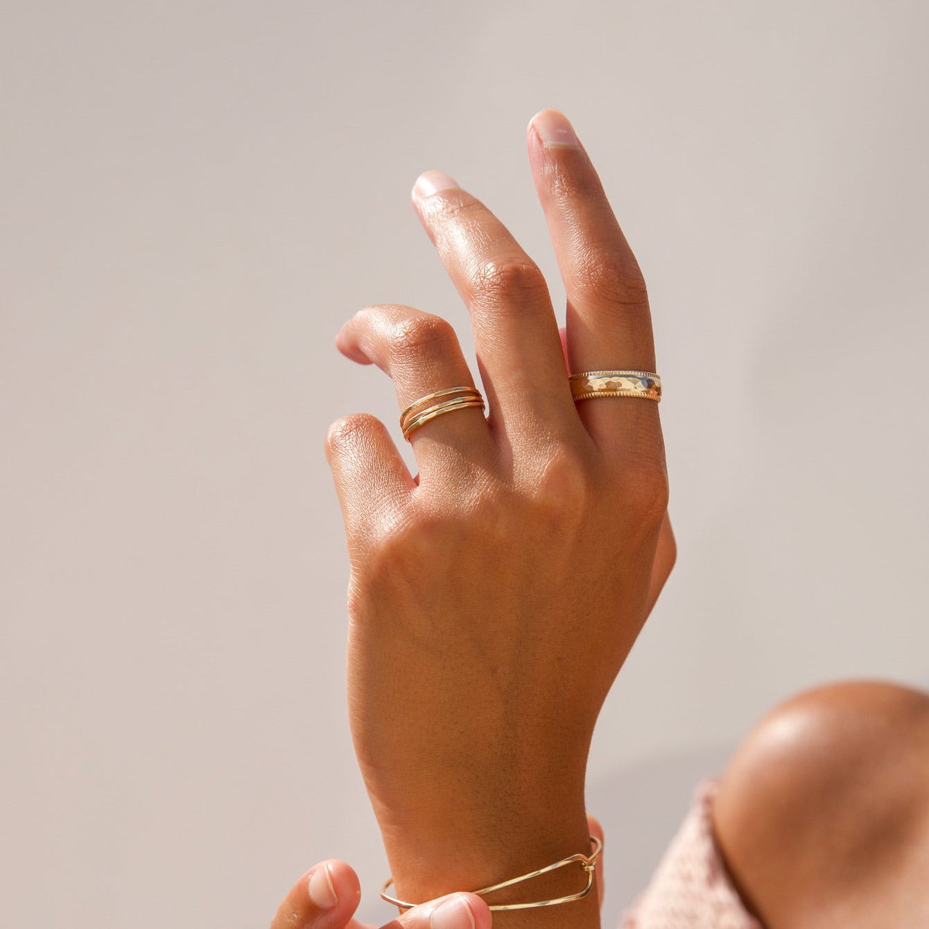 Close-up shot of a model wearing minimalist gold jewelry in warm sunlight, featuring stacked thin gold bands on the ring finger, a textured wide gold band on the pointer finger, and a sleek open cuff bracelet on the wrist.
