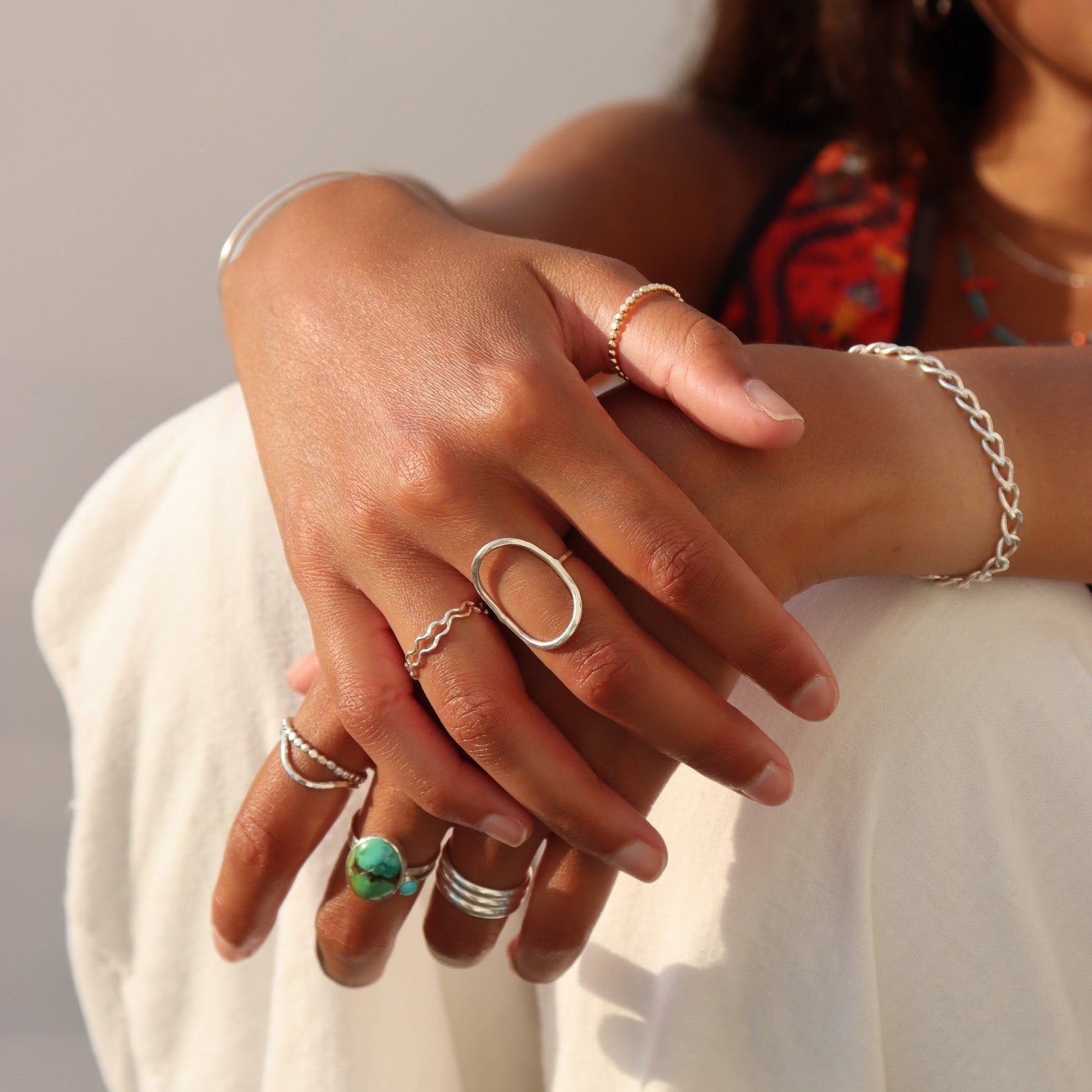 Close-up lifestyle image of a person wearing layered sterling silver rings, including a bold oval statement ring, wavy stacking ring, beaded bands, and a natural turquoise statement ring. A chunky silver chain bracelet and slim cuff complete the look, photographed in warm golden light against a soft neutral background.