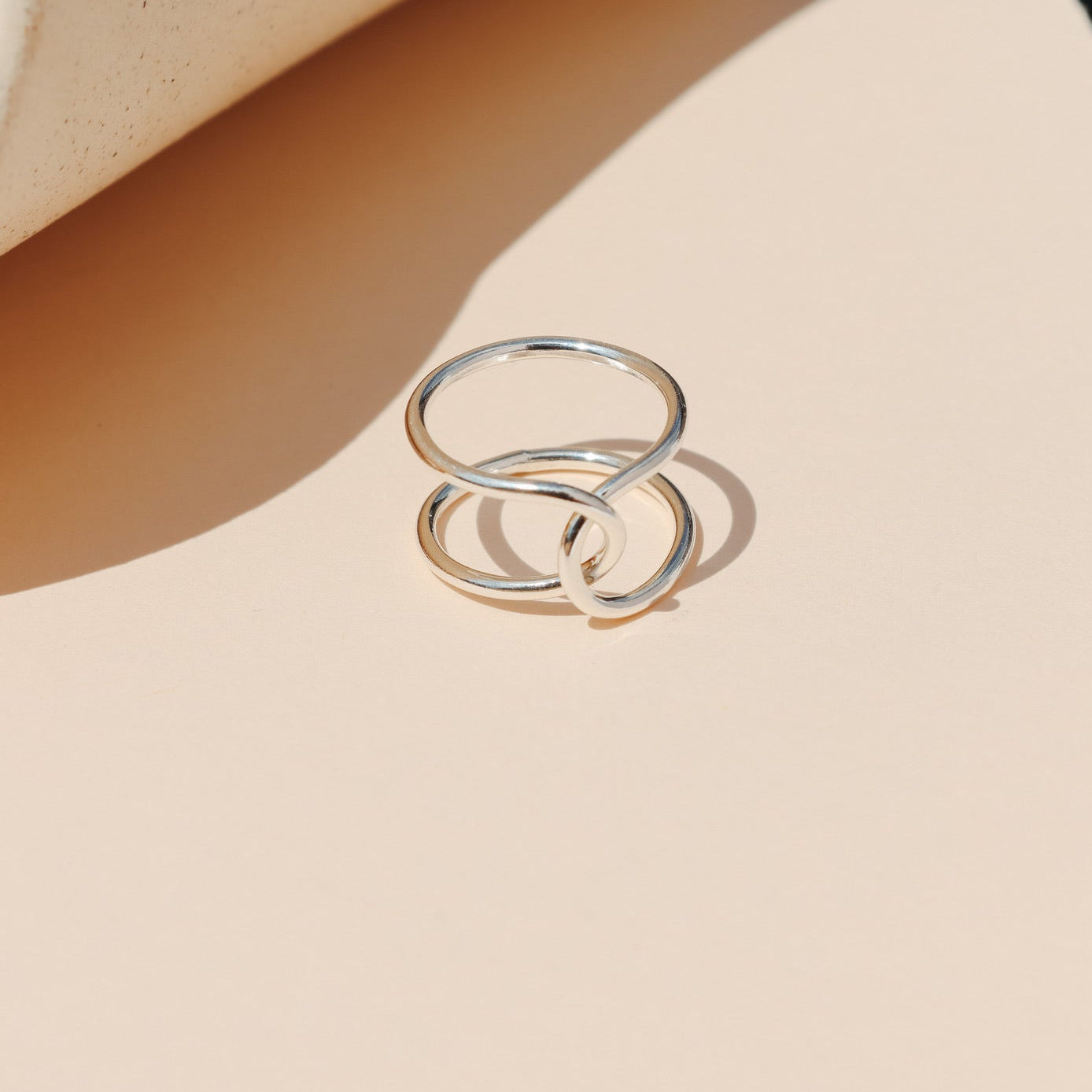 Minimalist sterling silver double-band ring featuring an intertwined loop structure, displayed on a warm peach-toned background with natural sunlight shadows.
