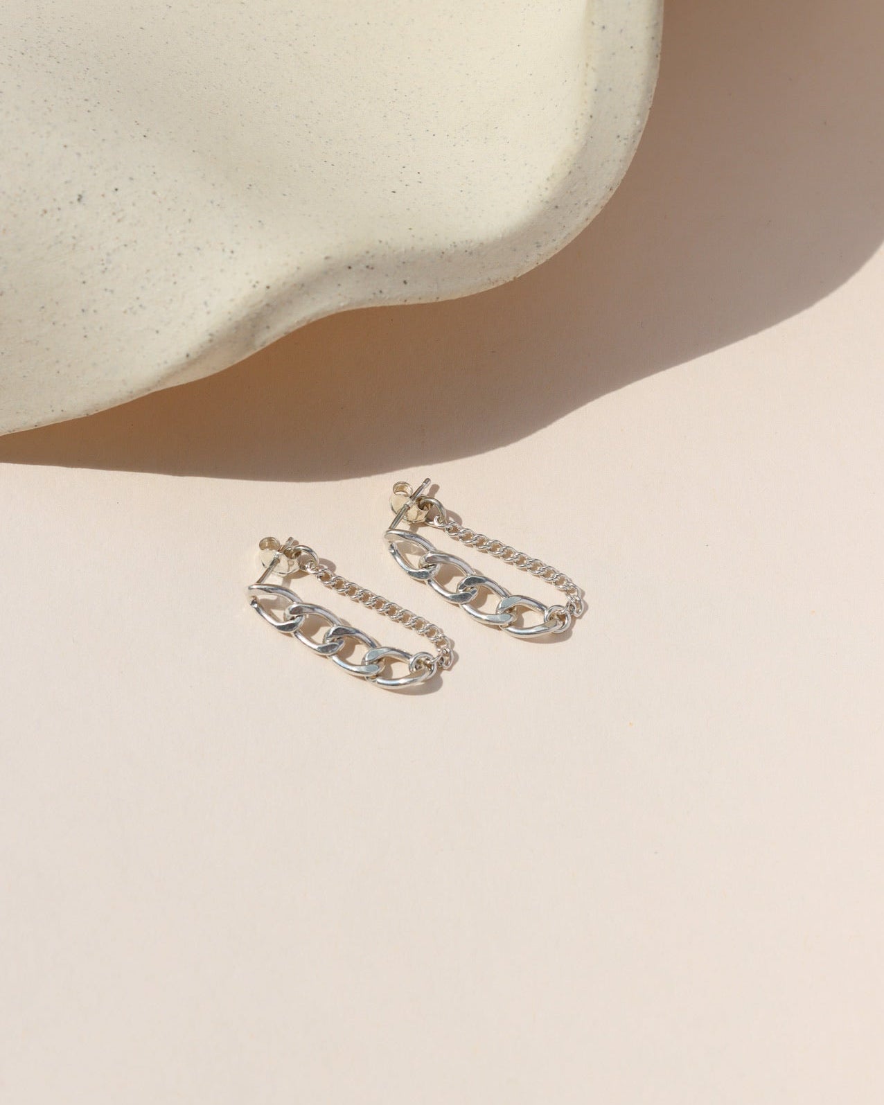 A pair of silver stud earrings featuring a dangling loop made of two combined chain styles—a chunky curb chain front and a fine link chain back. Laid flat on a light beige surface with soft shadows cast by a ceramic tray in bright sunlight.