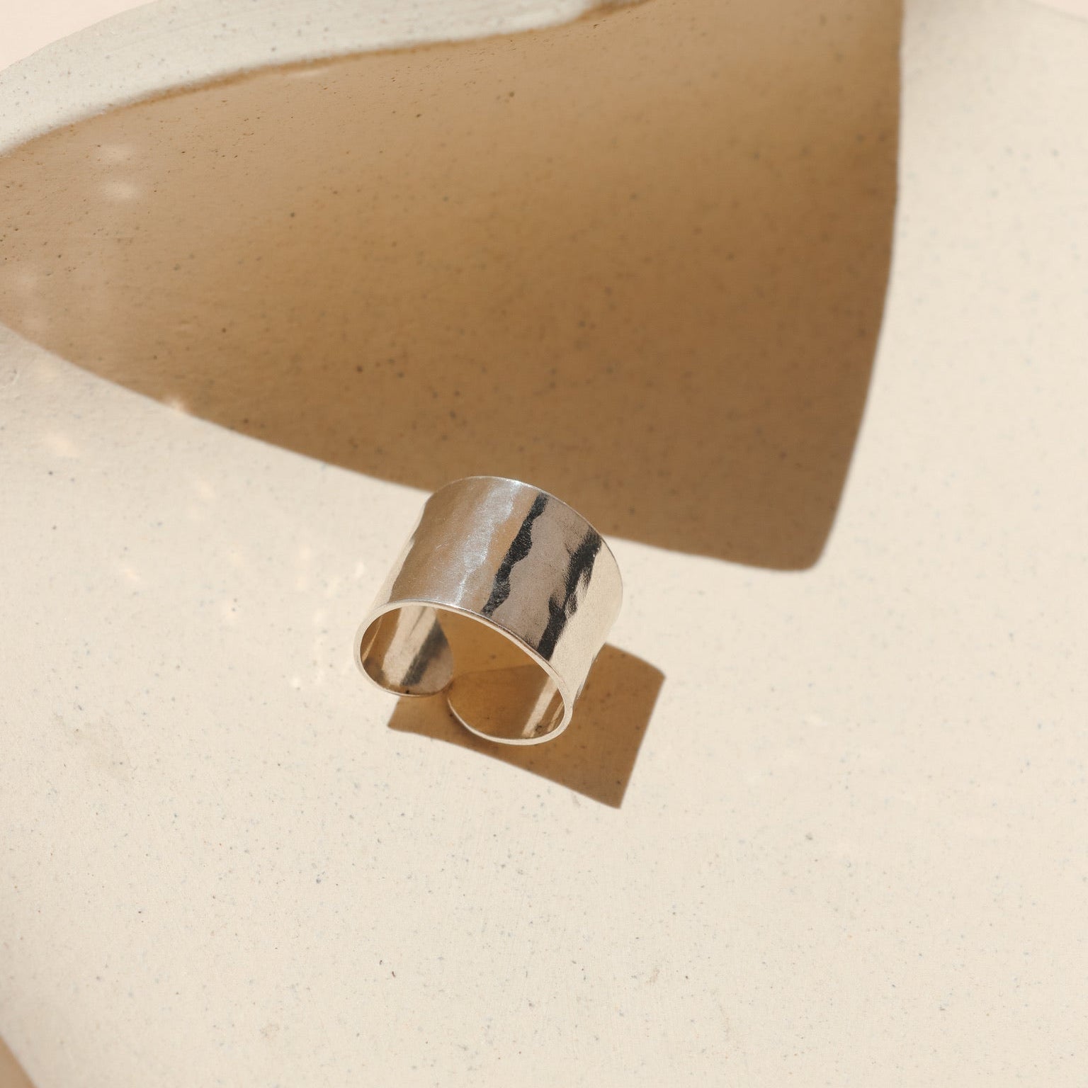 Wide sterling silver statement ring with a textured, hammered finish and an open adjustable back, set inside a neutral speckled ceramic prop with warm sunlight shadows.