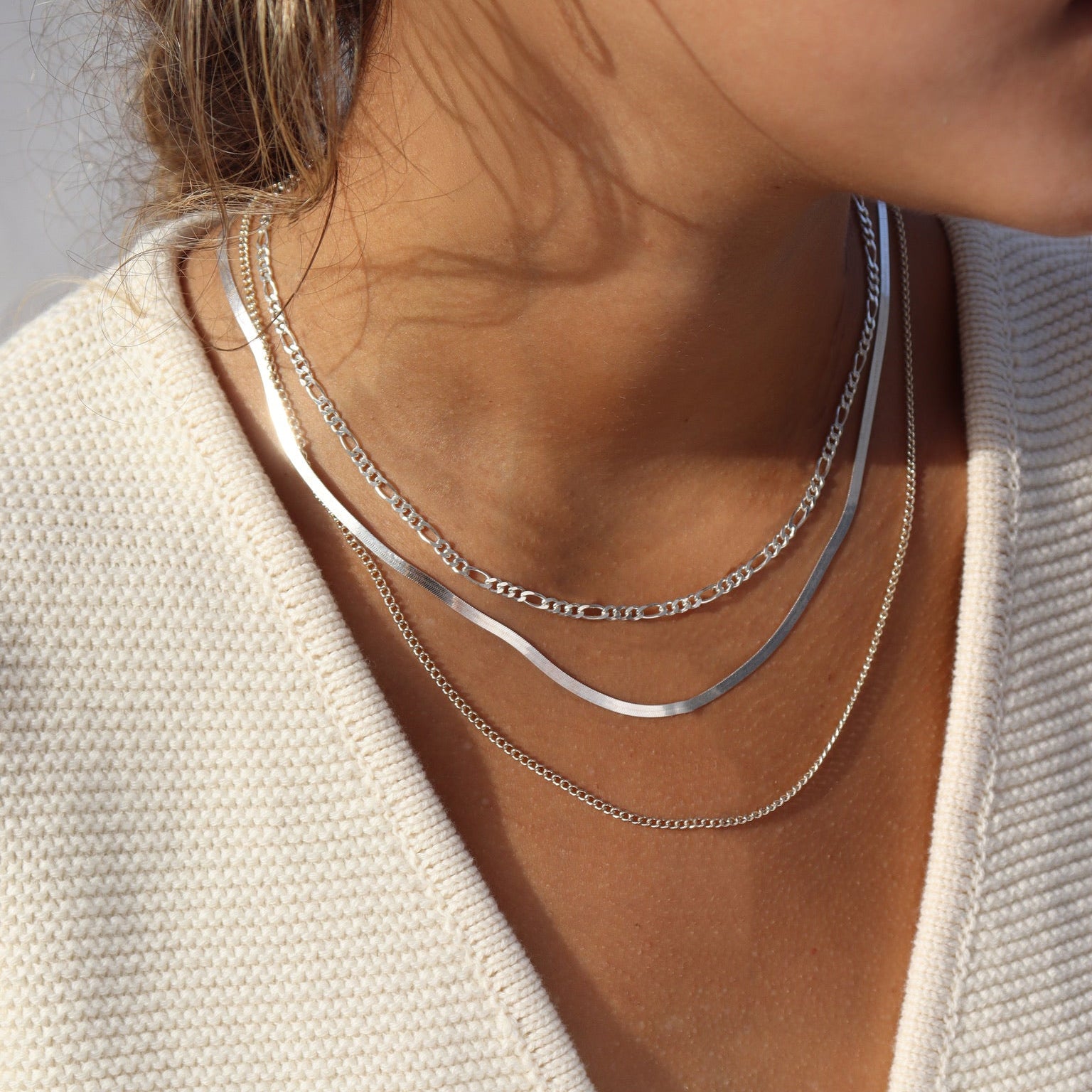 Close-up shot of a woman's neckline in warm natural sunlight, styling a three-tier sterling silver chain set: a top Figaro chain, a sleek middle herringbone snake chain, and a delicate bottom curb chain paired with a cream knit sweater.