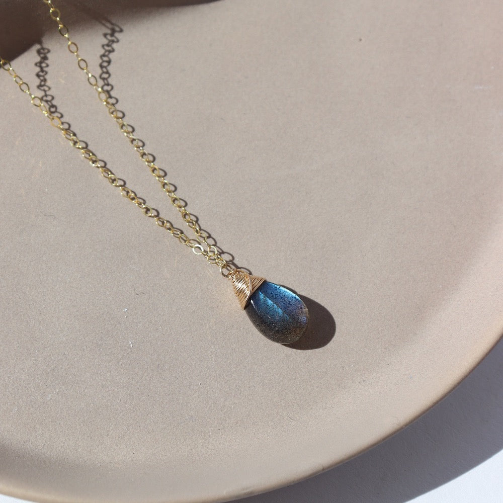 14k gold fill Stormi Necklace laid on a tan plate in the sunlight. This necklace features the simple chain with the Labradorite teardrop gemstone.