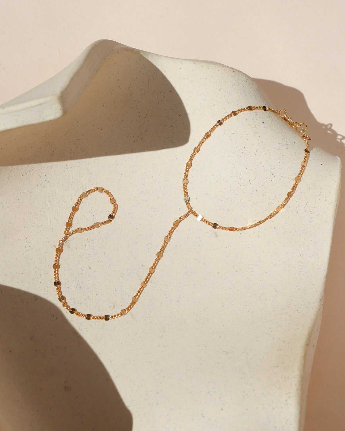Delicate gold hand chain with a fine textured chain connecting the bracelet to a finger loop, displayed on a cream ceramic form in warm sunlight.