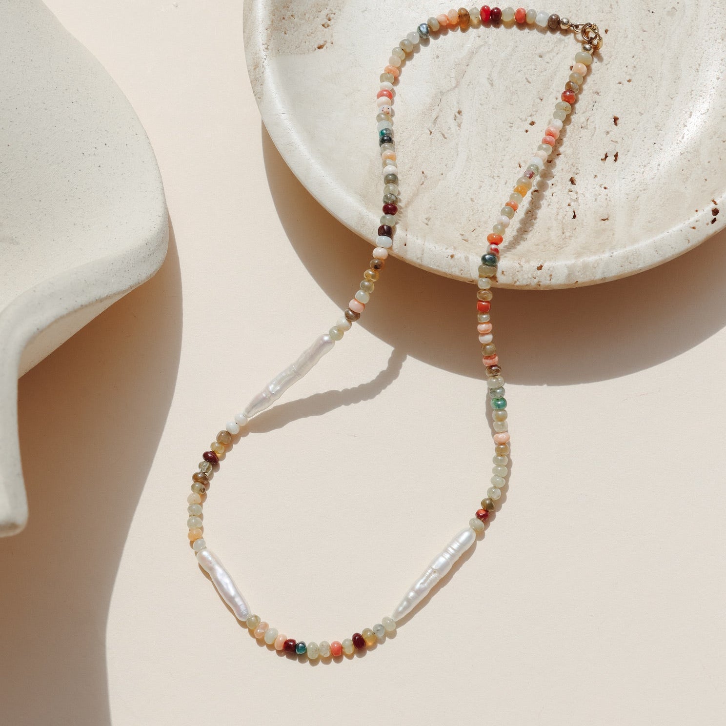 Multicolored Mexican Opal beaded necklace accented with three elongated freshwater pearls draped on a beige dish in the sunlight.