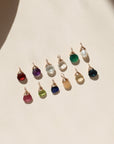 Assortment of colorful faceted gemstone charms in red, purple, pink, green, blue, opal, and pearl tones, each finished with gold wire wrapping, arranged on a cream background in warm sunlight.