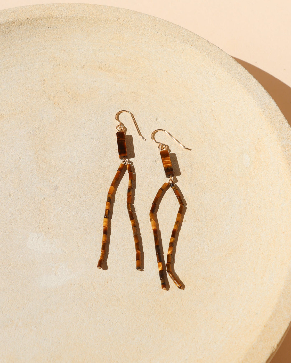 A pair of dangling earrings with gold filled french ear wires and rectangular Tiger's Eye gemstones and two long, Tiger's Eye beaded strands dangling, displayed on a textured beige ceramic dish.