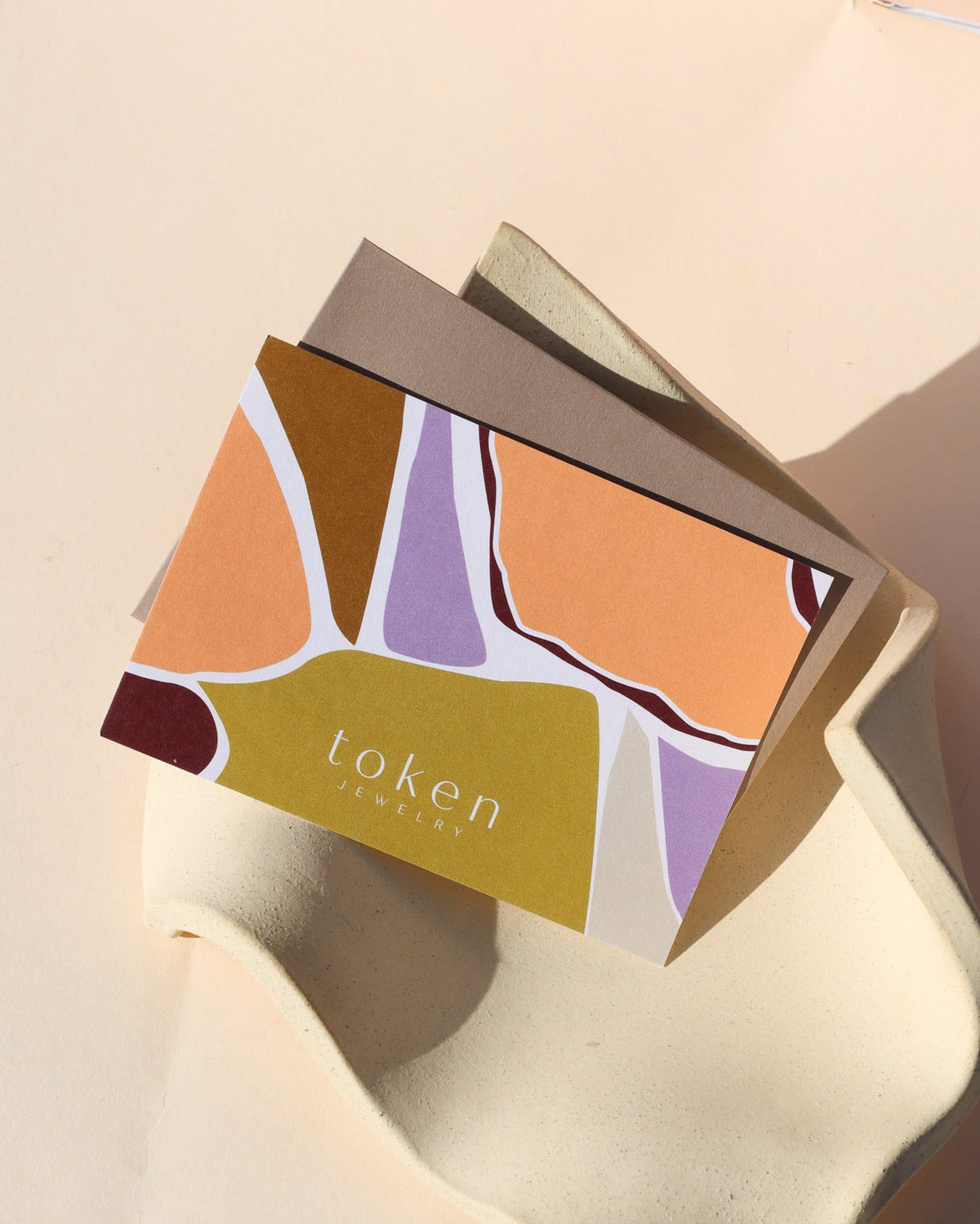 Token Jewelry gift card featuring an abstract design in warm orange, mustard yellow, lavender, and burgundy, displayed with a kraft envelope on a sculptural cream ceramic tray in soft sunlight.