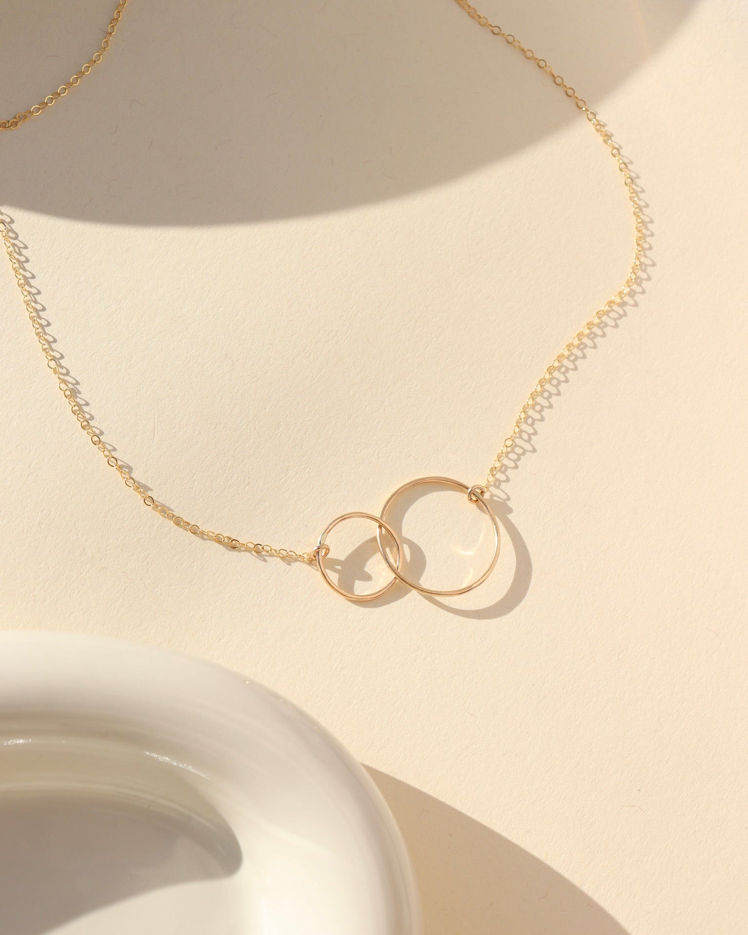 Top-down view of a fine gold chain necklace with an interlocking double-circle pendant, laid out on a cream-colored surface with warm natural sunlight casting subtle shadows next to a white ceramic dish