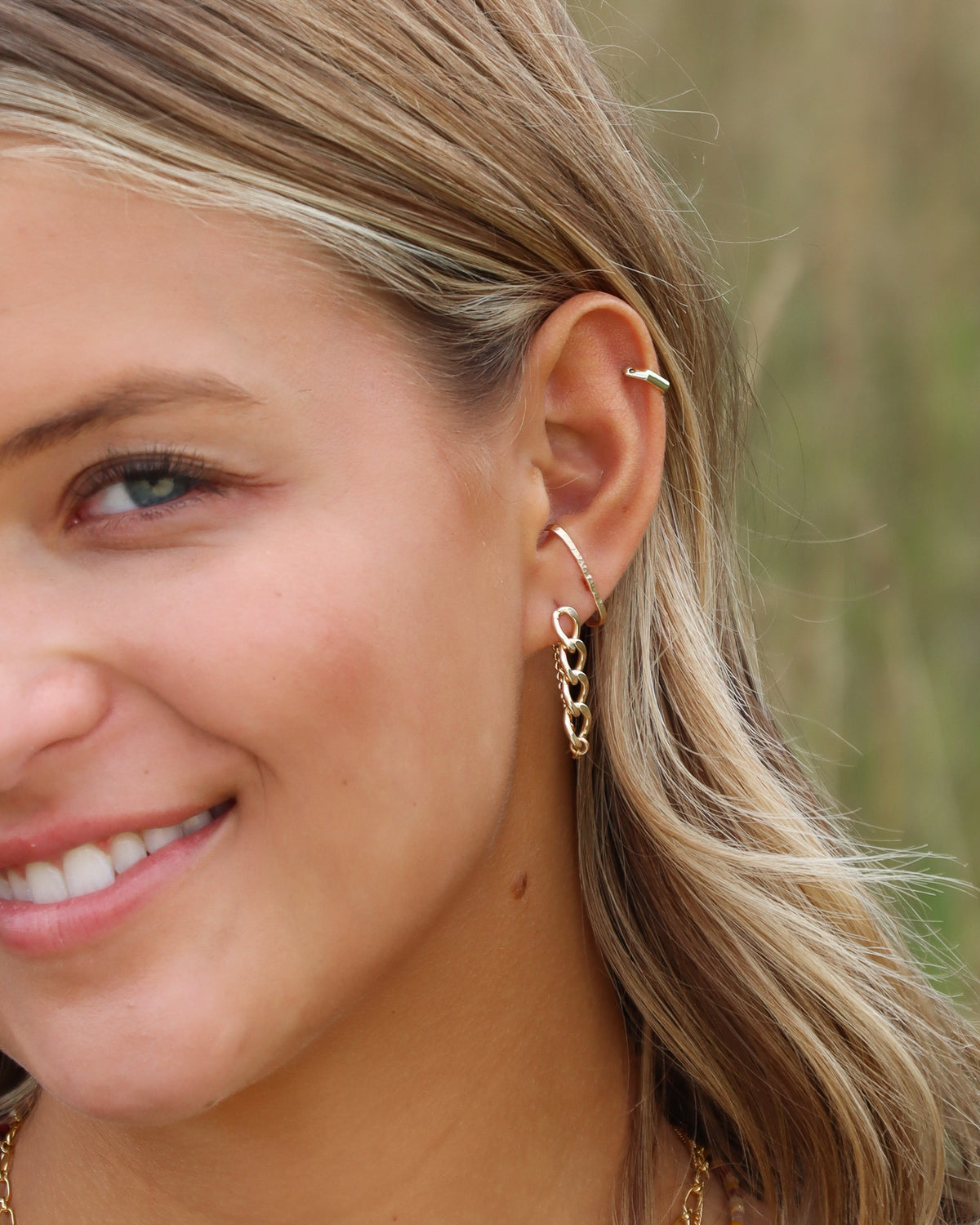 A close-up of a woman's ear wearing a gold ear cuff with a dangling chain of heart-shaped links and a small gold hoop piercing behind her ear, set against a blurred natural background.