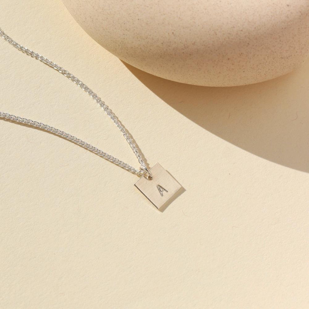 A small silver chain with a 3/8 x 3/8 square pendant stamped with a "G". Necklace is displayed on a cream backdrop. 