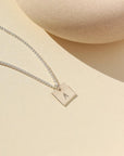 A small silver chain with a 3/8 x 3/8 square pendant stamped with a "G". Necklace is displayed on a cream backdrop. 