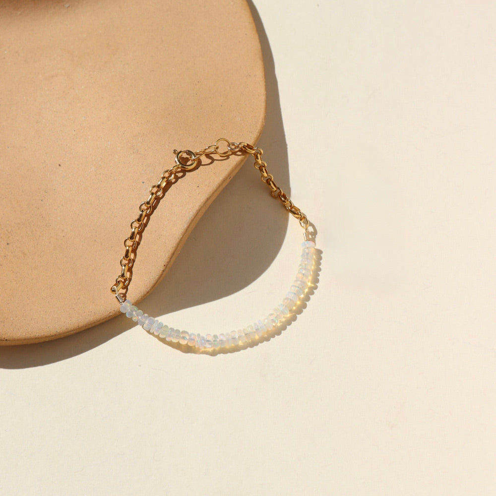 A 14k gold fill half and half bracelet featuring white opal gemstones and a rolo chain with a standard gold clasp. The bracelet is displayed on a cream backdrop in the sunlight. 