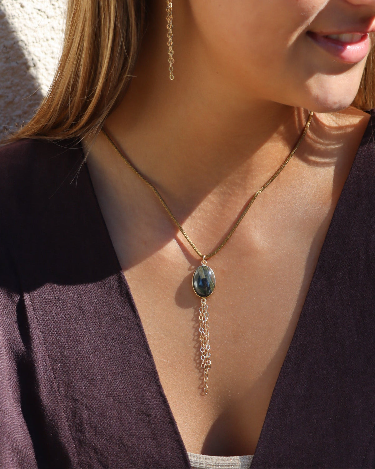 Oval dark multicolored gemstone pendant necklace with delicate chain fringe, styled with a brown blouse in warm sunlight.