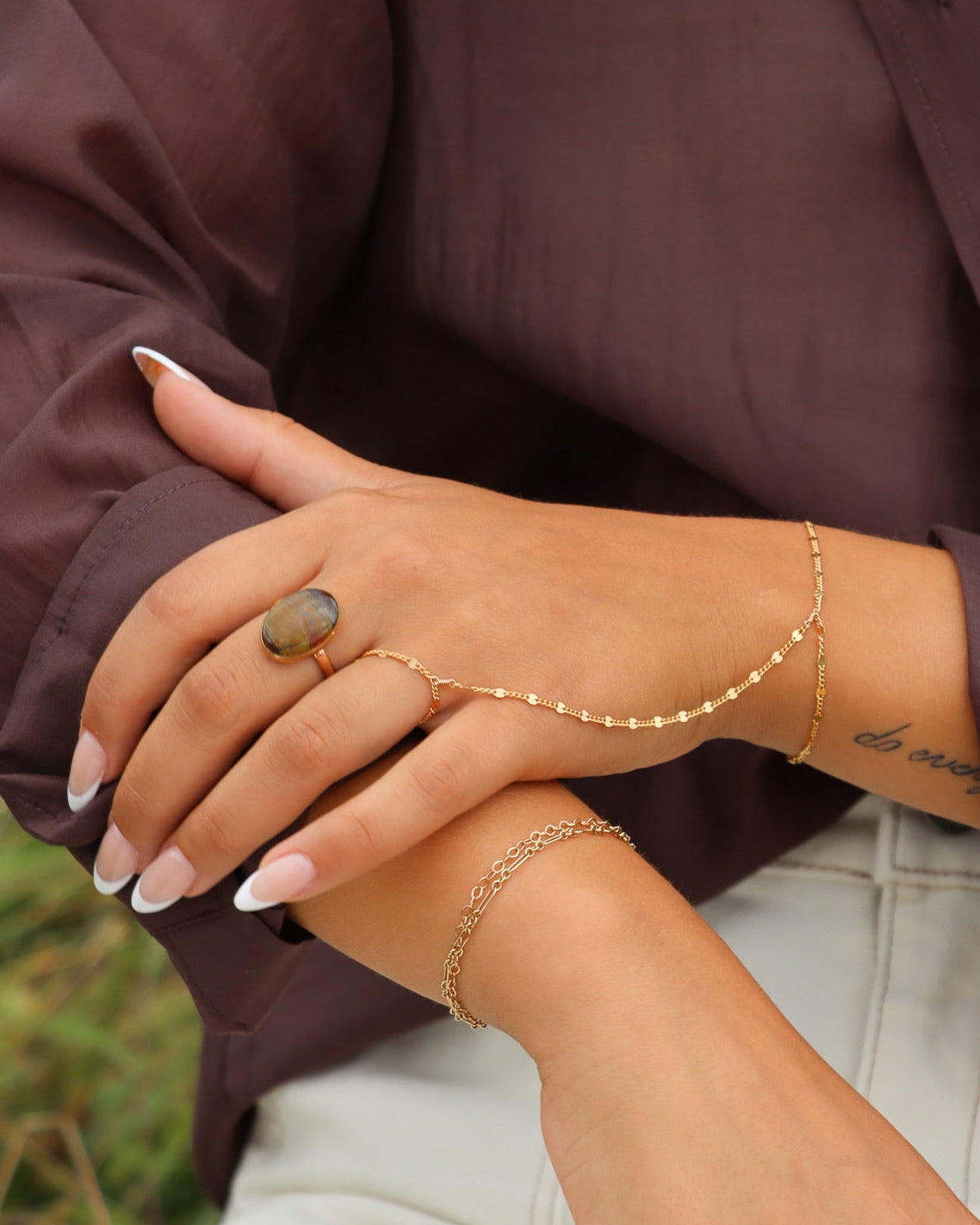 Close-up of layered gold bracelets, hand chain, and gemstone rings styled with a brown blouse and cream pants.