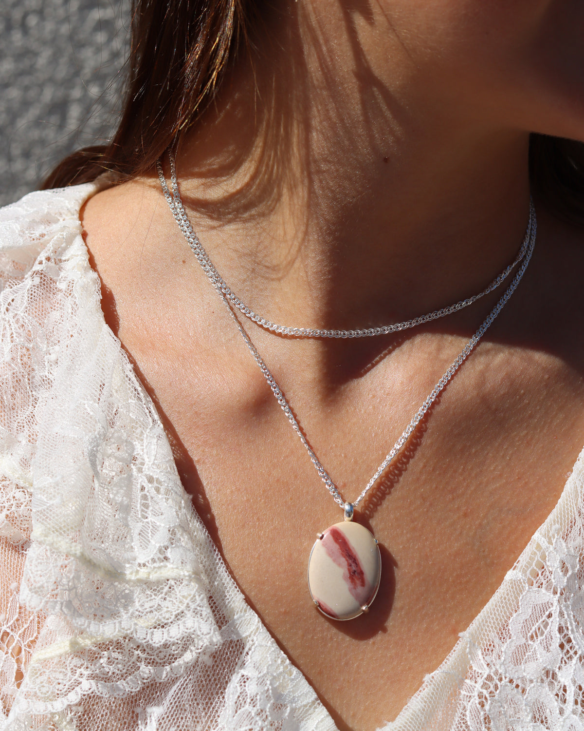 Layered sterling silver necklaces featuring a woven chain and an oval cream and red gemstone pendant, styled in warm sunlight.