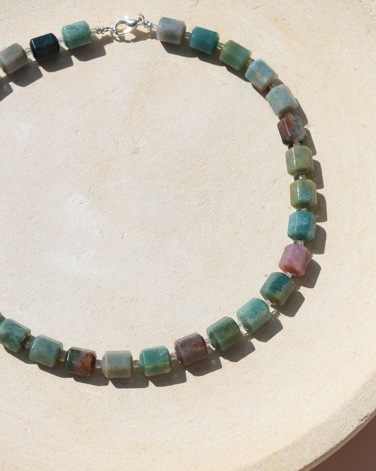 Beaded gemstone necklace featuring polished barrel-shaped stones in shades of green, teal, blue, and earthy pink, finished with a silver clasp and displayed on a textured cream surface in natural sunlight.