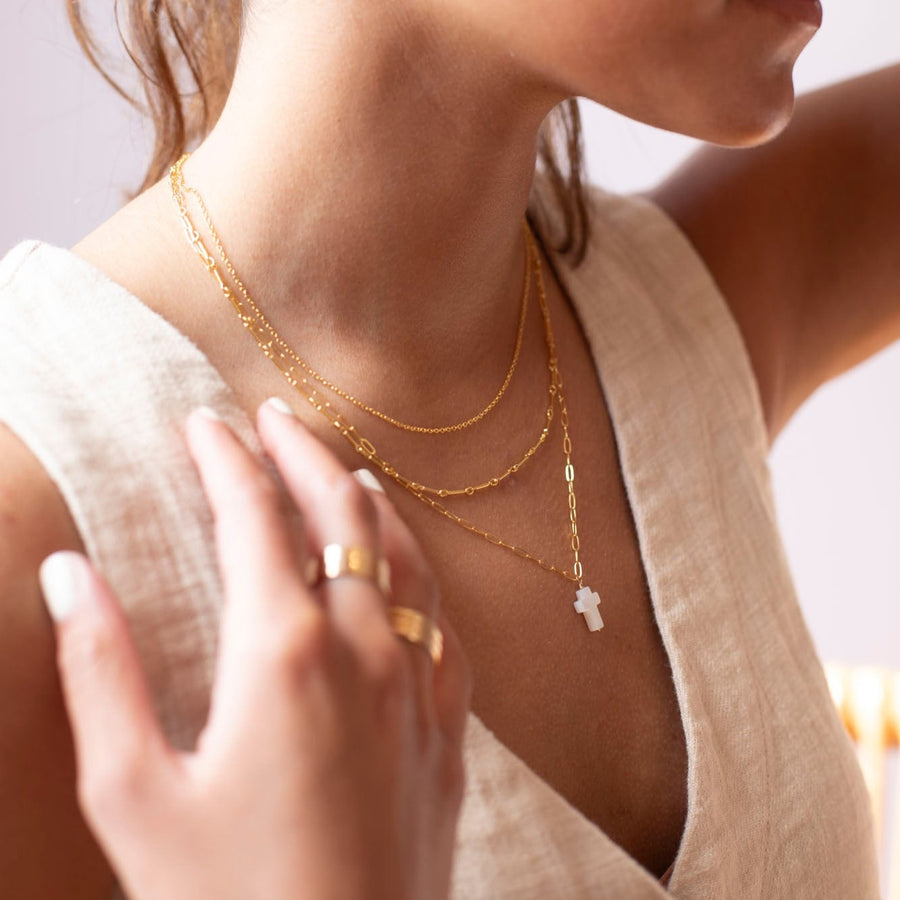 a mother of pearl white cross pendant on a 14k gold fill chain, photographed on a model wearing a cream linen vest and other 14k gold fill chain necklaces made by Token Jewelry