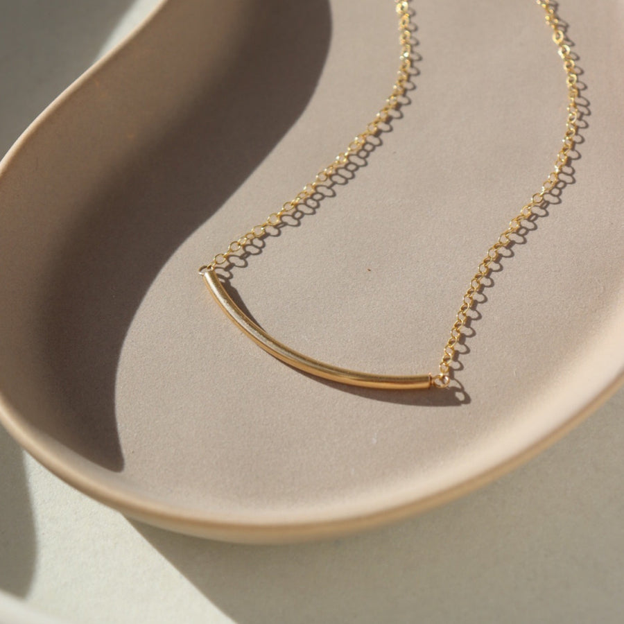 14k gold fill Minimal Necklace placed on a tan plate in the sunlight. This Necklace features a small bar about two inches long and connected by our simple chain.