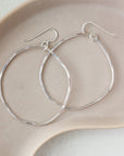 925 sterling silver form hoops placed on a peach plate set in the sunlight. These earring feature a large hoop that is hammered on both sides and has a hook for a earring