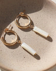 14k gold fill Mother of Pearl Hoops laid on a gray plate in the sunlight. These earring feature a classic hoop with a mother of pearl drop down stone.