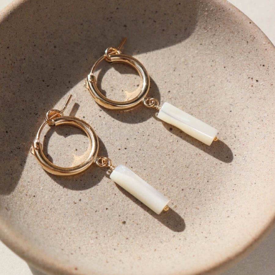 14k gold fill Mother of Pearl Hoops laid on a gray plate in the sunlight. These earring feature a classic hoop with a mother of pearl drop down stone.
