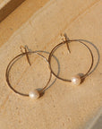 14k gold fill Pearl Hoops laid on a white plate in the sunlight. These earrings feature a hammered hoop with a pearl gemstone connecting the hoop together.
