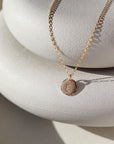 Small Monogram Coin Necklace laid on two white stone plates in the sunlight. This necklace features the coin chain followed by the mini coin tag that is hammered on the upside and is marked with the initial E.