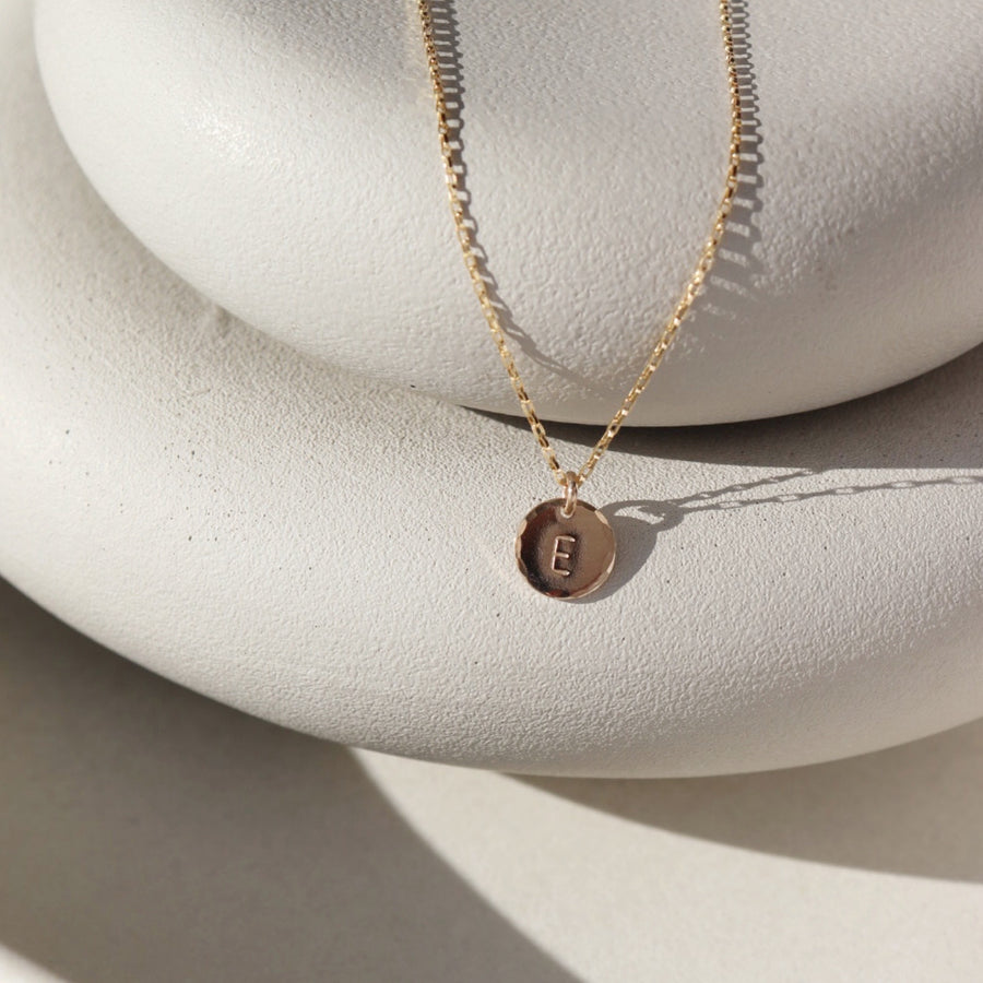 Small Monogram Coin Necklace laid on two white stone plates in the sunlight. This necklace features the coin chain followed by the mini coin tag that is hammered on the upside and is marked with the initial E.