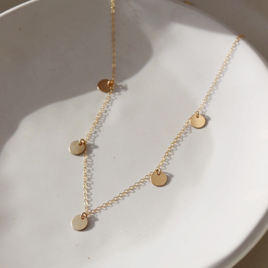 14k gold fill Suncatcher necklace laid on a white plate in the sunlight. This Necklace features the simple chain with 5 circle disc. This necklace is perfect for layering.