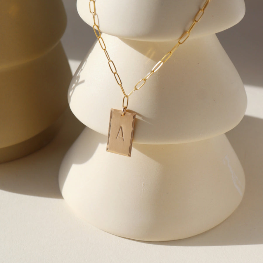 14k gold fill Everly Monogram Necklace placed on a white pot. Cosette linked chain - Gold or Sterling Silver Rectangle Hammered edge - Monogrammed with letter - Jewelry near me - Eau Claire