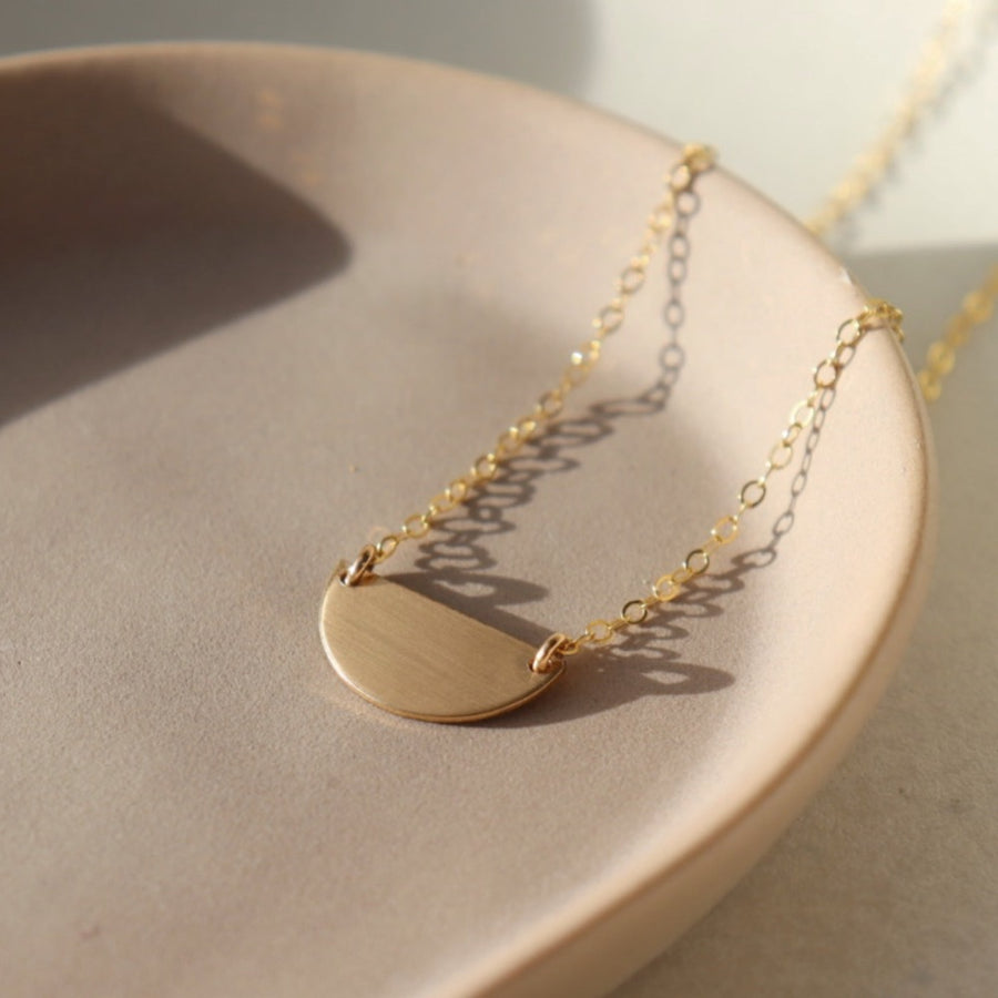14k gold fill Sunrise Necklace laid on a tan plate in the sunlight. This necklace features the simple chain and is connected by a half disc. The disc is smooth and gives the effect of a sunrise. 
