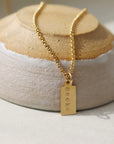 14k gold fill Mini Tag Personalized Necklace placed on a tan and gray bow. This necklace features the coin necklace chain followed by a mini tag with personalization of your choice.