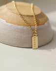 14k gold fill Mini Tag Personalized Necklace placed on a tan and gray bow. This necklace features the coin necklace chain followed by a mini tag with personalization of your choice.