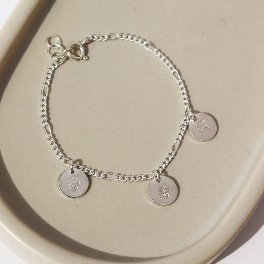 925 sterling silver Birth Flower Charm Bracelet laid on a tan plate in the sunlight. This Bracelet feature the Demi Alexandra chain with as many birth flower charms as you want.