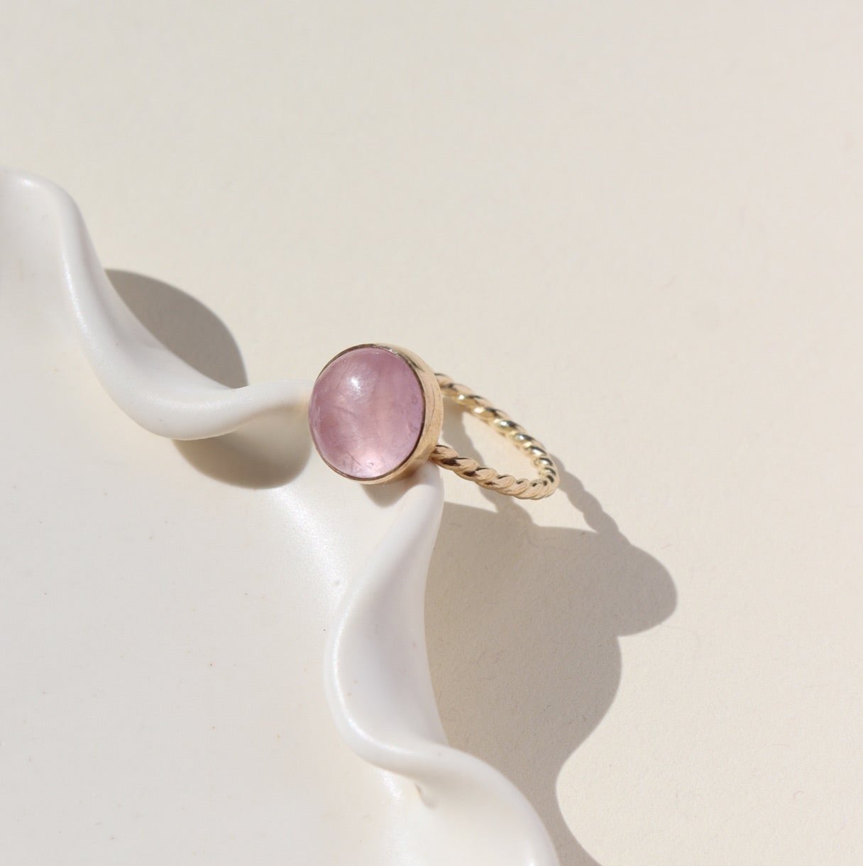 14k gold filled Pippa Ring laid on a white paper in the sunlight. This ring features a sprial band with a pink kunzite gemstone.