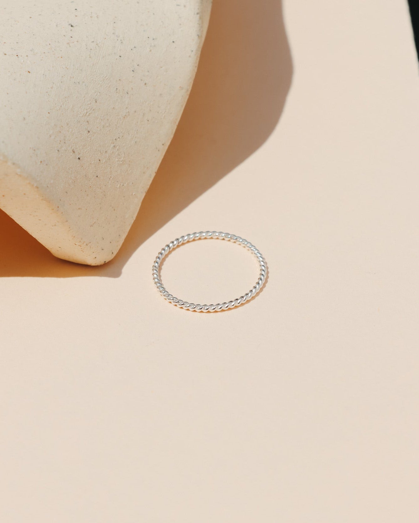 Minimalist sterling silver twisted rope stacking ring lying flat on a peach-beige background with a curved ceramic prop casting soft natural shadows