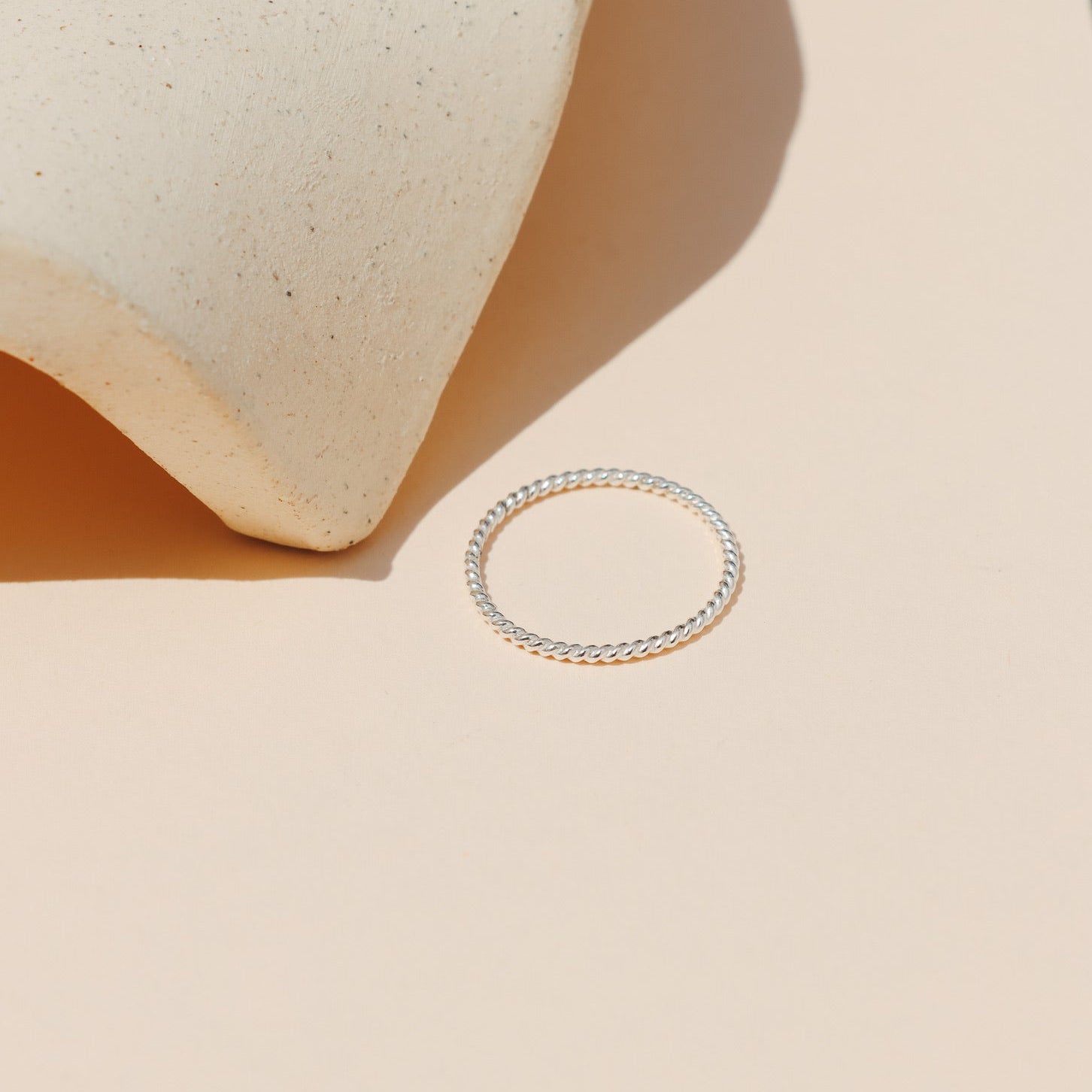 Minimalist sterling silver twisted rope stacking ring lying flat on a peach-beige background with a curved ceramic prop casting soft natural shadows