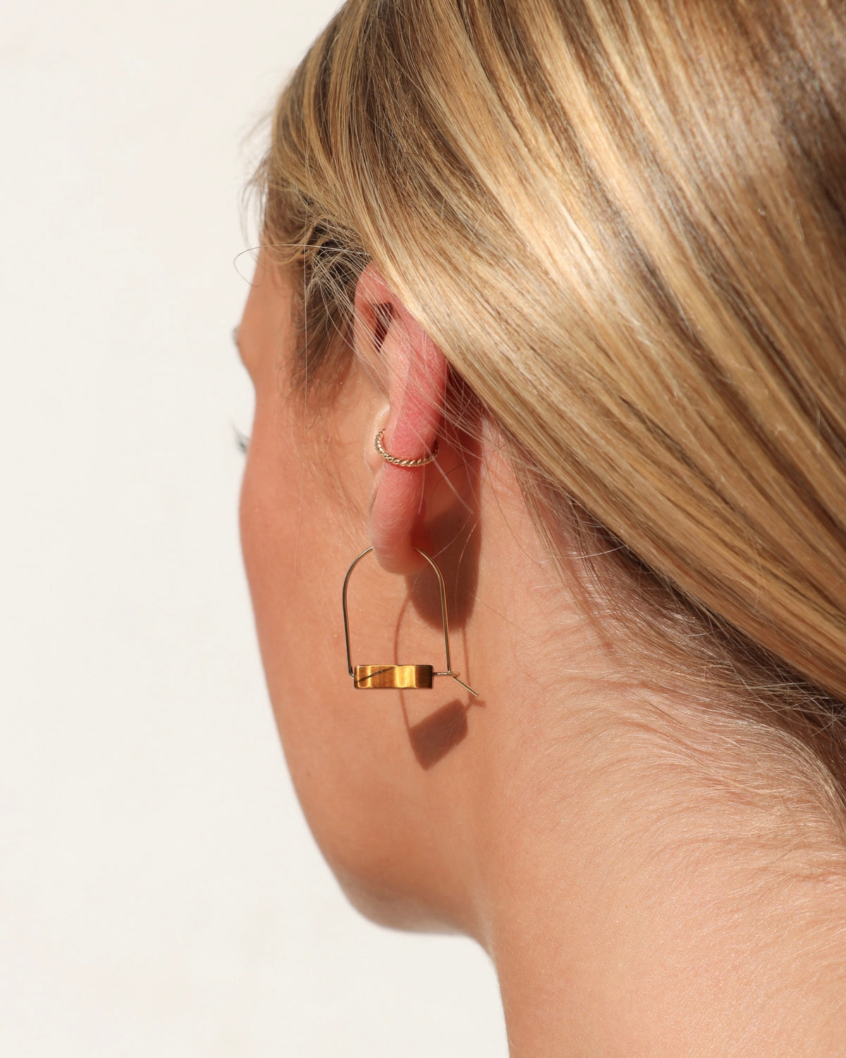 Model wearing gold wire drop earrings with warm brown Tiger’s Eye gemstone accents and a delicate gold ear cuff, photographed in natural sunlight against a soft white background.