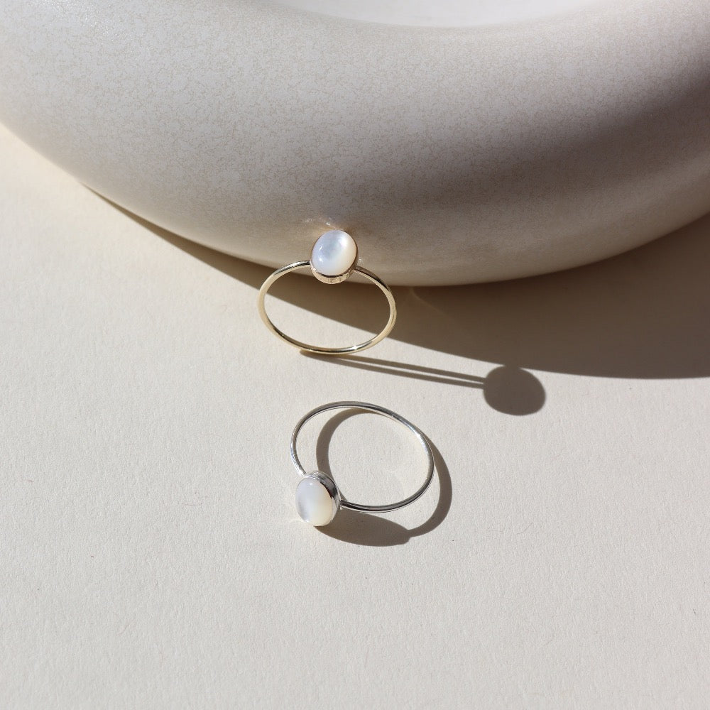 mother of pearl stone set in a bezel on a 14k gold fill skinny ring band, and a 925 sterling silver version, photographed on a table next to a white ceramic dish in the sun